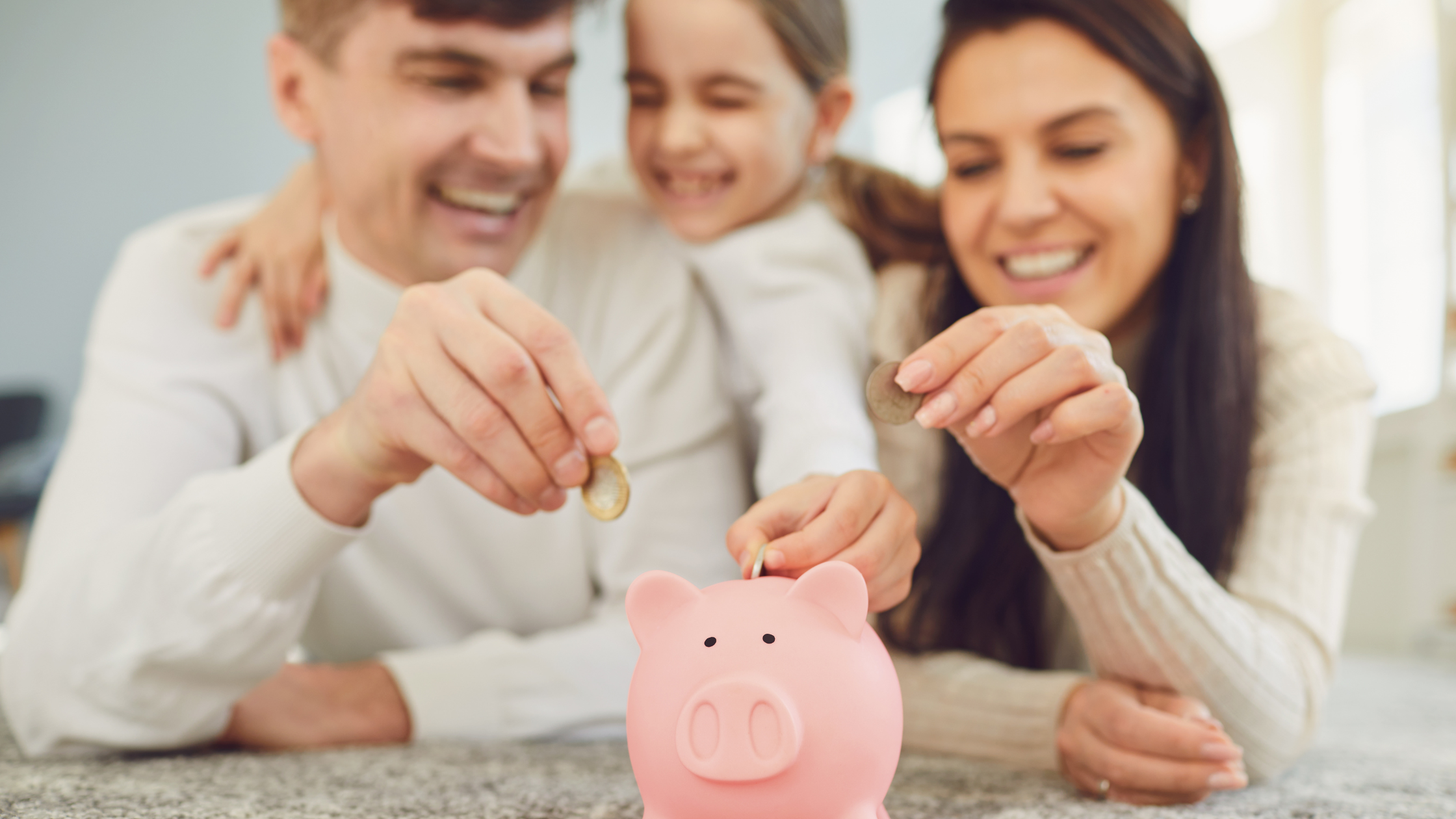 Portrait of happy little girl and parents sitting at kitchen table holding toy piggy bank.